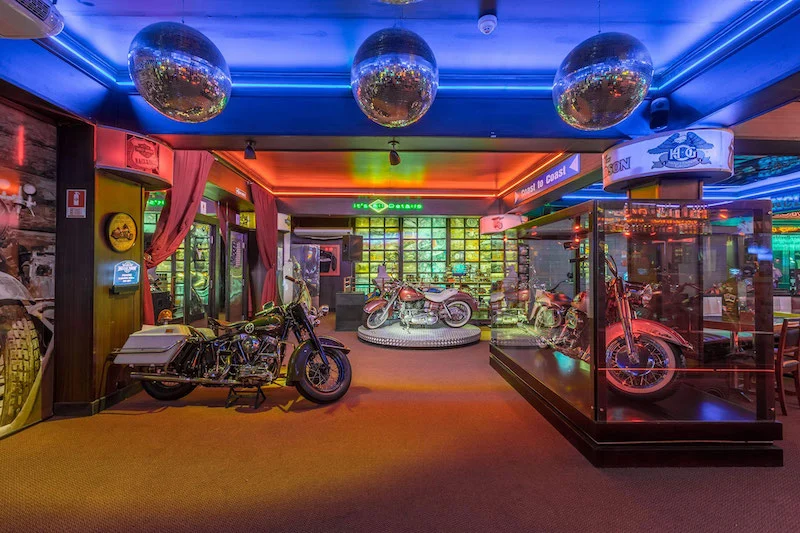 Ambiente interno do Dreams Motor Show em Gramado, com motocicletas clássicas expostas em pedestais e vitrines de vidro, iluminação colorida em tons de azul, vermelho e verde, e decoração temática que remete a um museu retrô de motos.