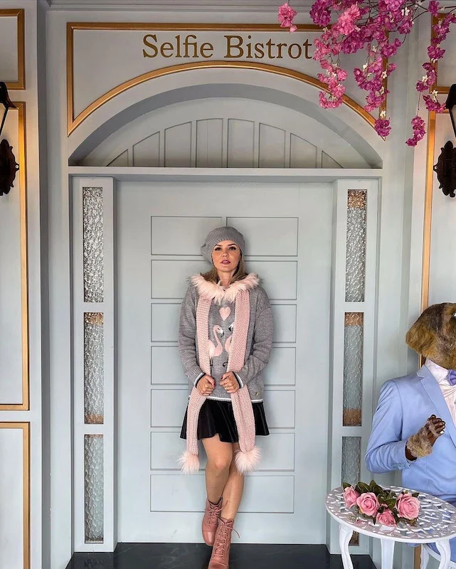 Visitante posa em um dos cenários temáticos do Selfie Gramado, em frente a uma fachada elegante com a inscrição ‘Selfie Bistrot’, usando roupas em tons pastel. O ambiente tem decoração charmosa, flores rosas suspensas e iluminação ideal para fotos criativas.