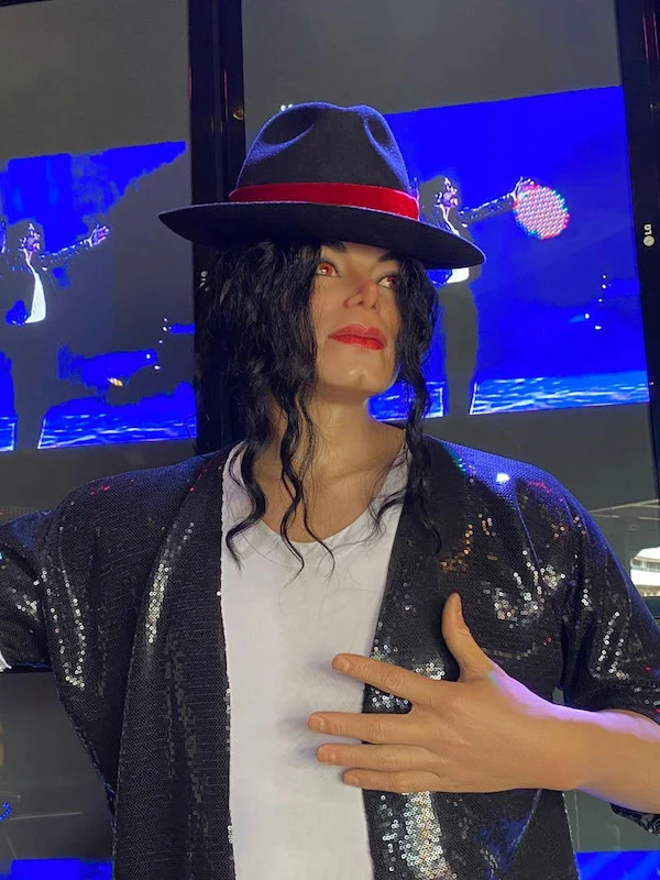 Réplica em cera de um cantor pop em pose de apresentação, vestindo chapéu preto, camisa branca e jaqueta com brilho, exposta no Museu de Cera de Gramado, com telões iluminados ao fundo.