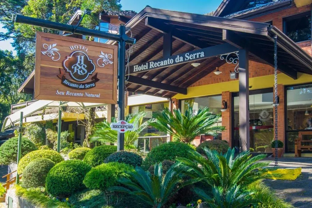 Fachada do Hotel Recanto da Serra, em Gramado (RS), com jardim bem cuidado, letreiro de madeira e entrada acolhedora sob um pergolado, em meio à vegetação típica da Serra Gaúcha.