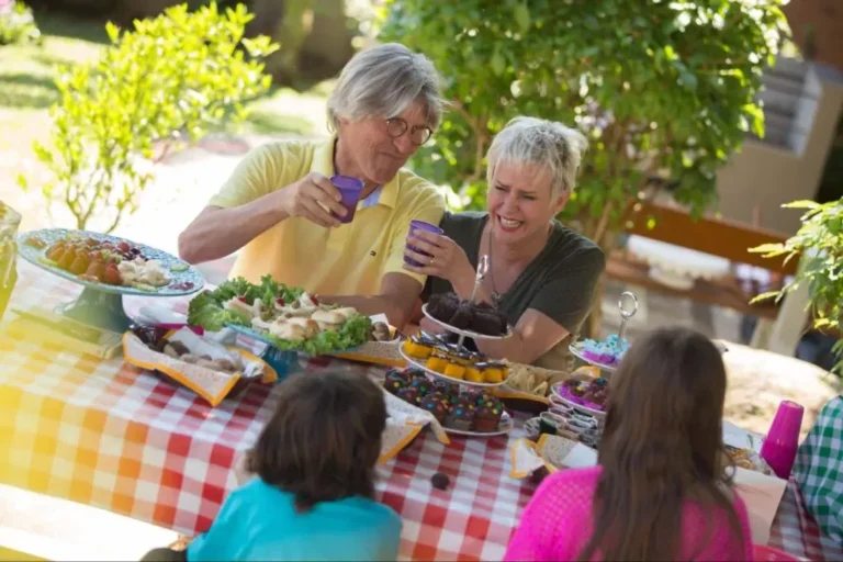 Grupo de pessoas sorrindo e brindando em uma mesa ao ar livre com toalha xadrez, repleta de comidas e sobremesas coloridas, em meio a um jardim ensolarado, representando momentos de lazer e convivência típicos de quem visita Gramado e está hospedado no Hotel Recanto da Serra.