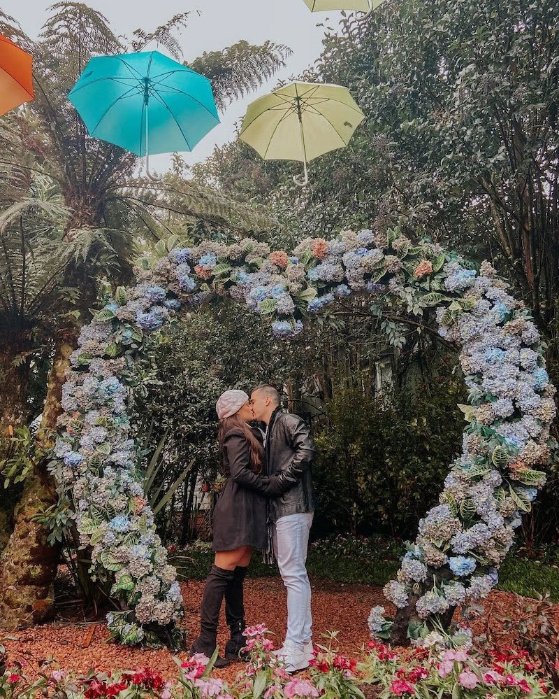 Casal se beijando sob um arco de flores em formato de coração no Jardim do Amor, representando o clima romântico dos parques em Gramado e Canela, com guarda-chuvas coloridos suspensos ao fundo.