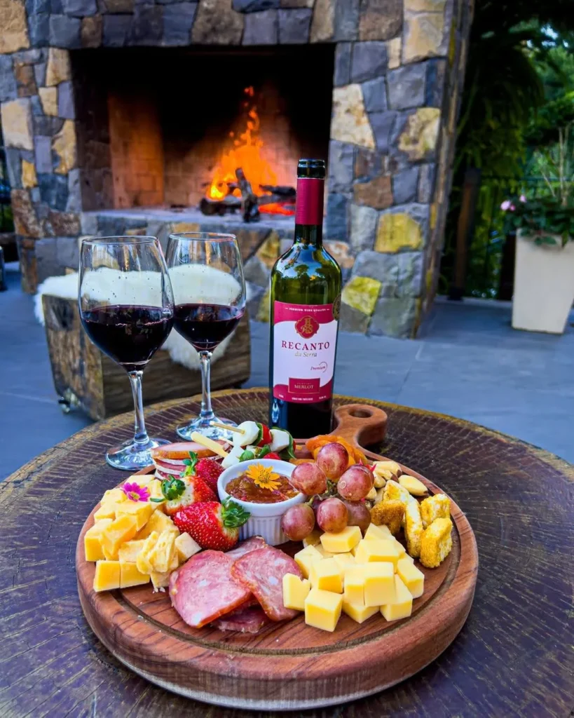 Taças de vinho tinto e uma garrafa de vinho Recanto da Serra ao lado de uma tábua de frios com queijos, embutidos, uvas, morangos e petiscos, acompanhados de uma geleia em um pequeno recipiente. A cena é iluminada pela luz do fogo de uma lareira a lenha, criando um ambiente acolhedor e sofisticado. Um cenário perfeito para desfrutar de uma refeição no hotel Centro de Gramado Recanto da Serra.
