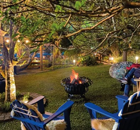 Ambiente aconchegante ao ar livre, com duas cadeiras azuis ao redor de uma fogueira, iluminada por luzes suaves de árvores e lanternas no jardim. A cena transmite um clima acolhedor e tranquilo, perfeito para momentos de relaxamento no hotel Centro de Gramado Recanto da Serra.