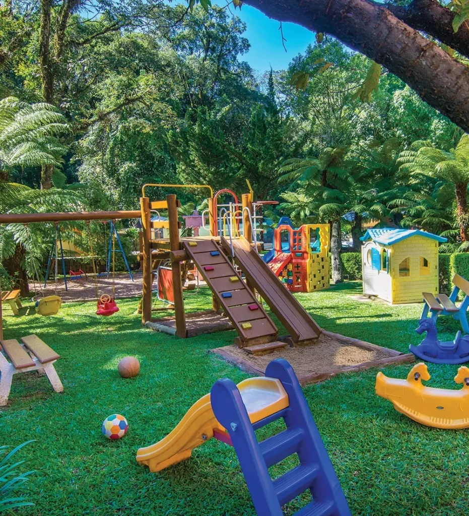 Área de lazer infantil no hotel em Gramado, com brinquedos coloridos, como escorregadores, balanços e casinha, rodeados por uma paisagem verde e árvores. O espaço ao ar livre oferece um ambiente seguro e divertido para as crianças aproveitarem durante a estadia.