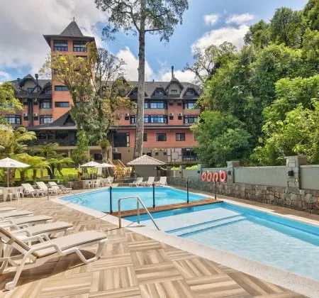 Piscina ao ar livre com espreguiçadeiras ao redor, localizada no hotel no centro de Gramado. O edifício do hotel, com sua arquitetura charmosa e cercado por vegetação, está ao fundo, com árvores e um céu claro ao fundo, criando um ambiente relaxante e convidativo para os hóspedes.