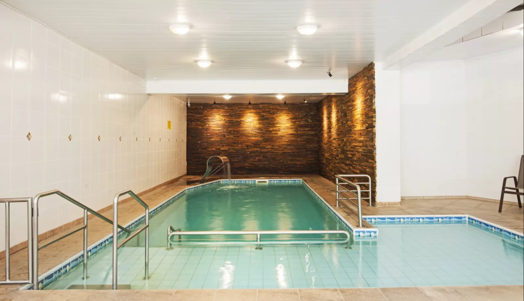 Piscina interna aquecida com escada metálica e um belo painel de pedras ao fundo, criando um ambiente relaxante e acolhedor. As luzes suaves iluminam a área, proporcionando uma experiência tranquila para os hóspedes do hotel no centro de Gramado.