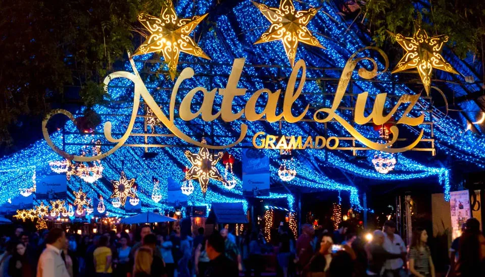 Fachada iluminada com o letreiro “Natal Luz Gramado” cercado por luzes azuis e estrelas douradas, com pessoas passeando e apreciando o evento noturno nas ruas da cidade.