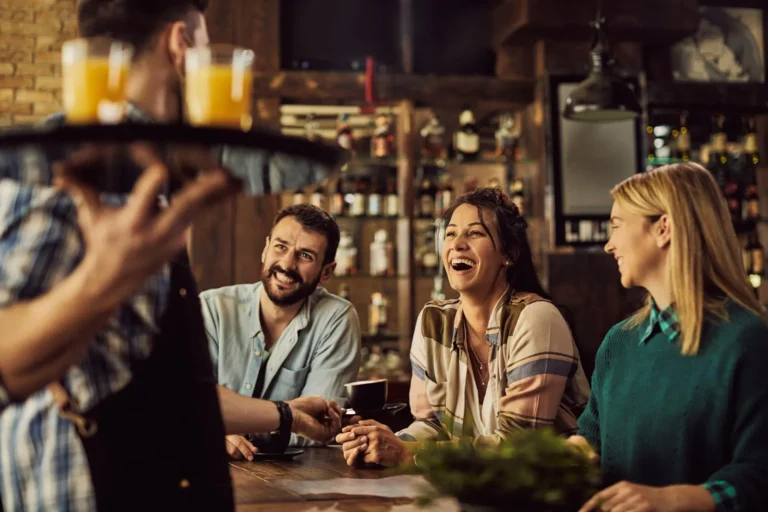 Grupo de amigos sorrindo e conversando em um restaurante aconchegante, com bebidas sendo servidas por um garçom, representando o clima acolhedor e gastronômico de Gramado.