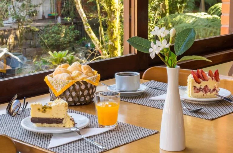 Mesa de café da manhã em ambiente aconchegante com vista para a natureza, com cesta de pães, fatias de torta, suco de laranja e café, representando o charme das cafeterias em Gramado.