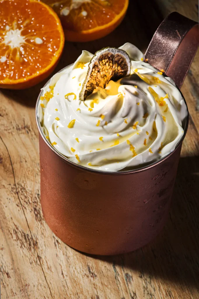 Imagem de um drink servido no Queen's Pub, localizado no Hotel Recanto da Serra, em Gramado. O drink é decorado com chantilly, raspas de laranja e uma fatia de laranja desidratada, representando a sofisticação e sabor dos bares em Gramado.
