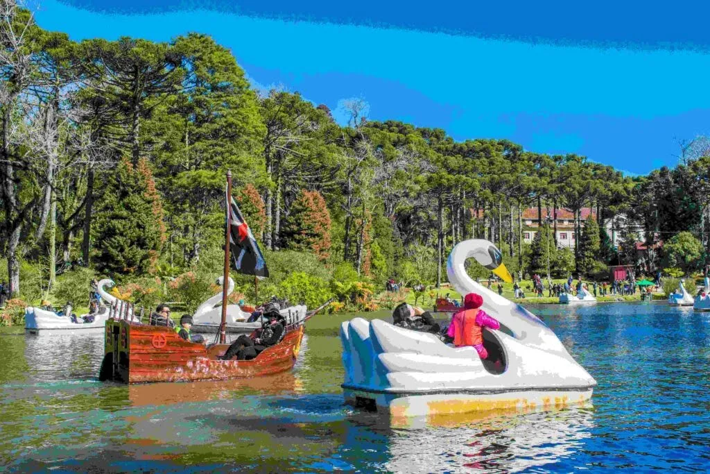 Pessoas se divertem em pedalinhos em formato de cisne no Lago Negro, em Gramado (RS), em um dia ensolarado. Entre os pedalinhos, há também um barco estilizado como navio pirata com bandeira preta. O lago é cercado por árvores altas e vegetação exuberante, com visitantes caminhando e apreciando a paisagem ao fundo.