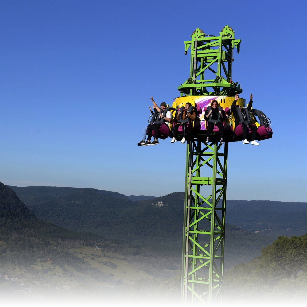 Brinquedo radical no Alpen Park em Canela, com visitantes sentados no topo de uma torre verde, suspensos sobre o vale e montanhas da Serra Gaúcha ao fundo sob céu azul.