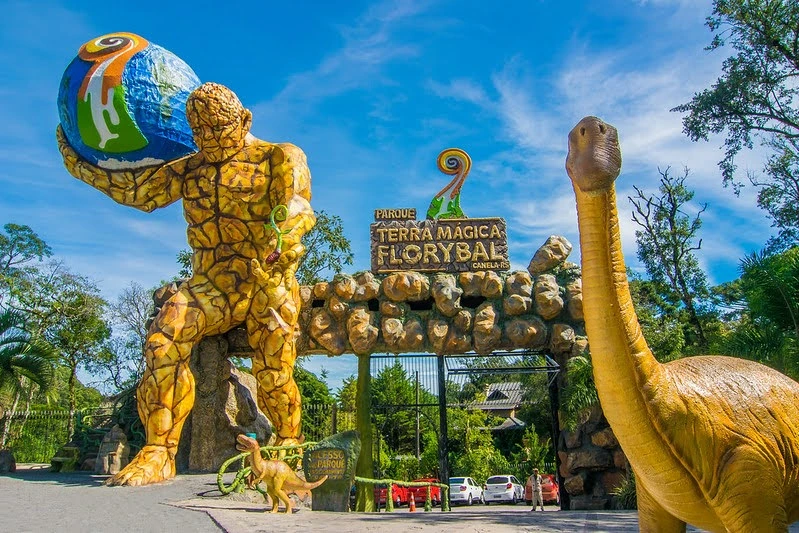 Entrada do Parque Terra Mágica Florybal em Canela, com esculturas gigantes de personagem segurando o planeta e dinossauro ao lado, portal temático de pedra e céu azul ao fundo.