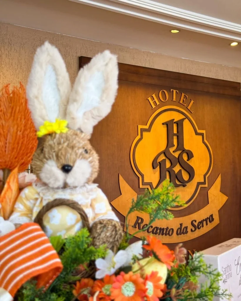 Decoração temática de Páscoa no interior do Hotel Recanto da Serra, com coelho decorativo, cenouras e flores coloridas em destaque. Ao fundo, placa de madeira com o nome do hotel, compondo um ambiente acolhedor e festivo que integra a programação de Páscoa 2026 em Gramado.