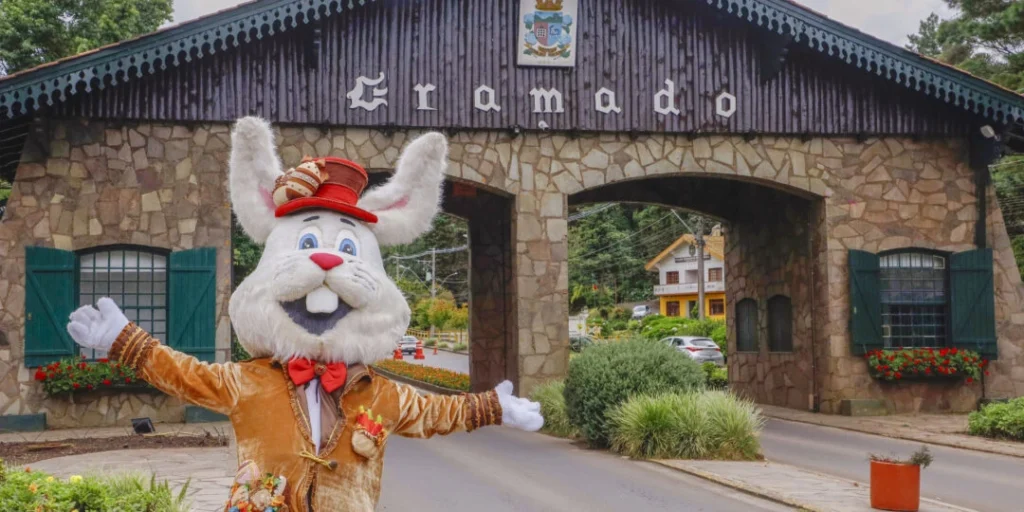 Personagem de coelho da Choco Páscoa em Gramado, vestido com traje elegante e cartola, posa com os braços abertos em frente ao pórtico de entrada da cidade. Ao fundo, o tradicional portal de pedra com a inscrição “Gramado” e arquitetura típica, cercado por vegetação, reforça o clima acolhedor e turístico do destino durante o evento.