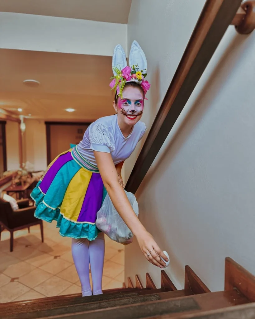 Funcionária caracterizada como coelhinha de Páscoa no Hotel Recanto da Serra, usando orelhas, maquiagem temática e roupa colorida, enquanto esconde ovos decorativos em uma escada. A cena representa a programação especial de Páscoa 2026 no hotel, com atividades lúdicas e interativas para os hóspedes.