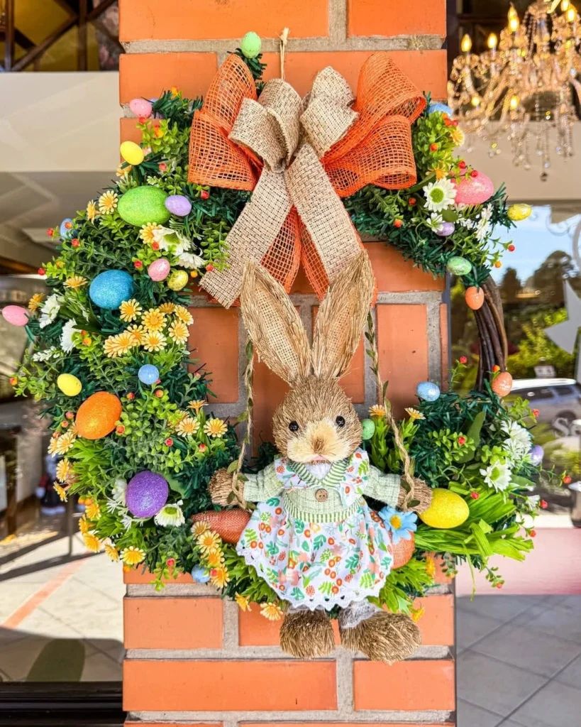 Guirlanda temática de Páscoa com coelho decorativo ao centro, cercada por flores, ovos coloridos e laços em tons naturais e laranja, fixada em uma parede de tijolos. A decoração representa o clima festivo da programação de Páscoa 2026 em Gramado, com elementos lúdicos e acolhedores.