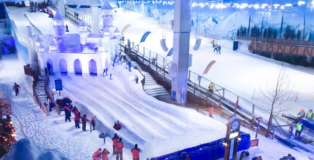 Parque Snowland em Gramado com pista de neve indoor, pessoas praticando esportes de inverno e descendo em boias, castelo cenográfico iluminado e ambiente temático que simula uma vila alpina.