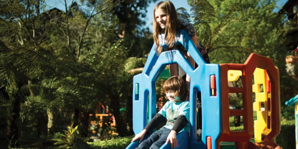 Espaço Kids do hotel com crianças se divertindo em brinquedo colorido ao ar livre, cercado por jardins e muita área verde, em um ambiente seguro, alegre e integrado à natureza.
