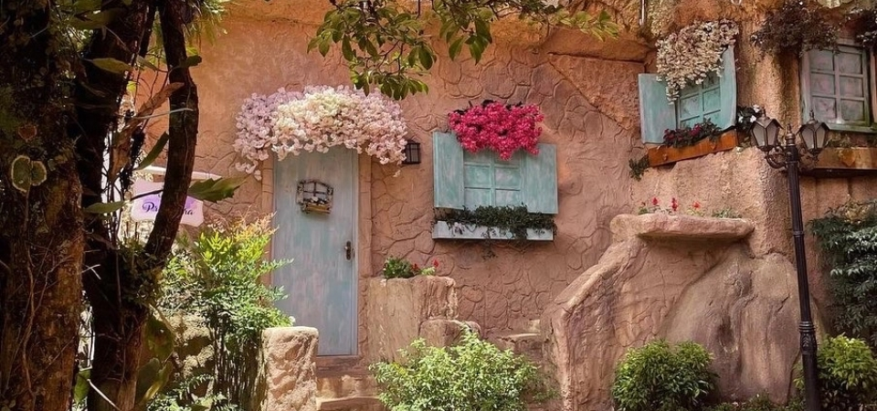 Fachada charmosa com flores e janelas coloridas em meio à natureza, representando o clima romântico do Jardim do Amor em Gramado.