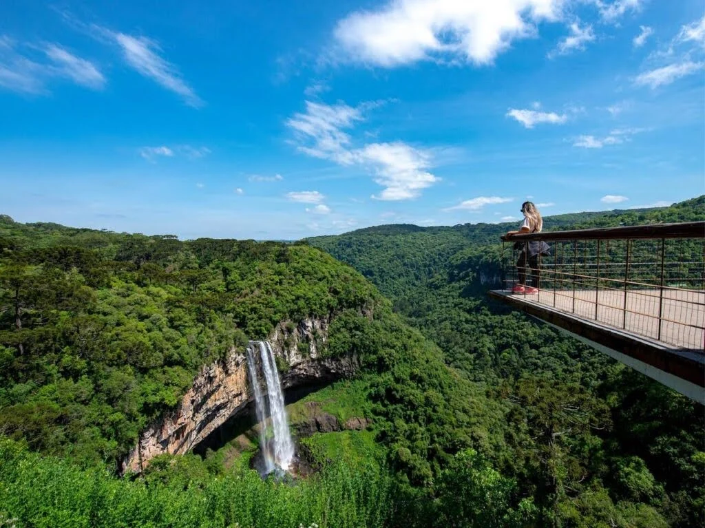 Mirante panorâmico no Parque Estadual do Caracol, em Canela (RS), com vista para a Cascata do Caracol, uma queda d’água imponente que despenca de um paredão de rochas cercado por densa vegetação de mata atlântica. Uma visitante observa a paisagem de cima da plataforma suspensa sob um céu azul com poucas nuvens.