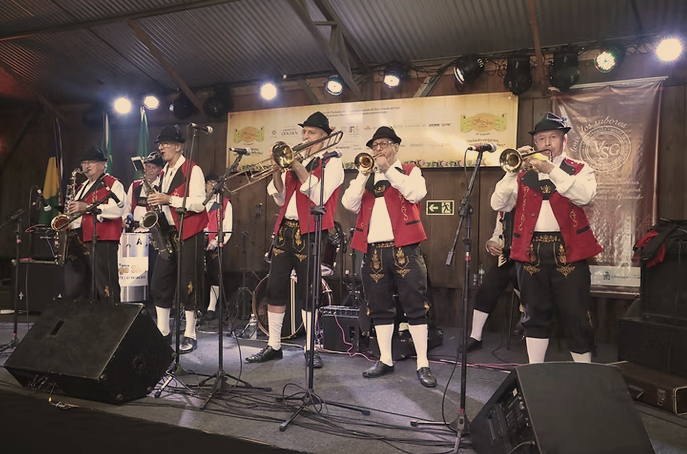 Banda tradicional se apresenta ao vivo em um palco coberto representando os eventos em Gramado. Os músicos vestem trajes típicos alemães com coletes vermelhos, chapéus e meias brancas, tocando instrumentos de sopro como trompetes e saxofones. A iluminação de palco e banners ao fundo sugerem um ambiente festivo e cultural.