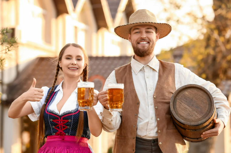Casal vestido com trajes típicos segurando canecas de cerveja e um barril, celebrando a Oktoberfest ao ar livre.