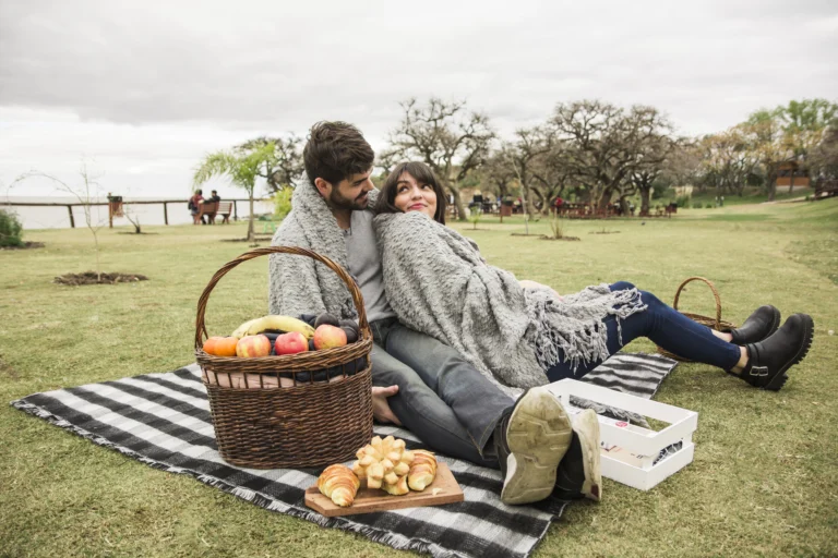Casal curtindo um piquenique romântico em um gramado, enrolados em um cobertor, com cesta de frutas e pães, inspiração perfeita para quem busca lugares para viagem a dois.