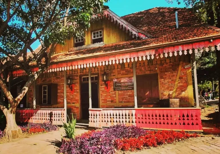 Fachada da Casa do Colono em Gramado, com arquitetura rústica em tijolos aparentes, detalhes de madeira e jardim florido em frente, localizada na charmosa Praça das Etnias.