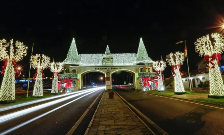 Pórtico de entrada de Gramado iluminado com luzes natalinas brancas e árvores decoradas com laços vermelhos, criando um cenário mágico durante as festividades de fim de ano.