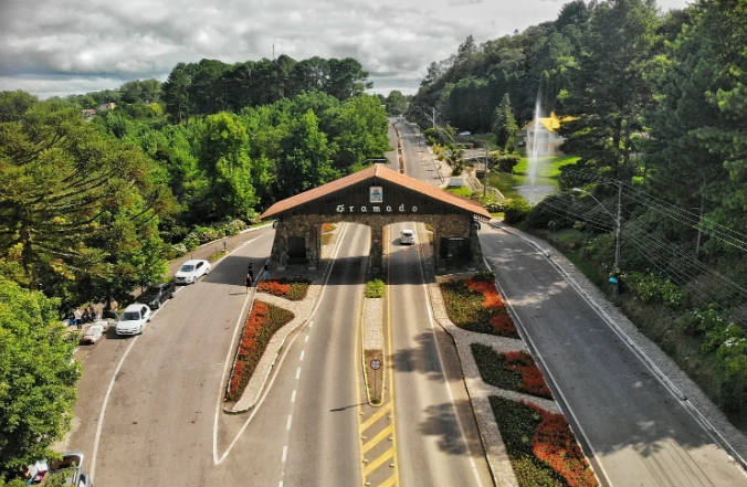 Vista aérea do pórtico de entrada de Gramado, cercado por jardins floridos, árvores verdes e vias bem cuidadas, marcando a chegada à charmosa cidade da Serra Gaúcha.