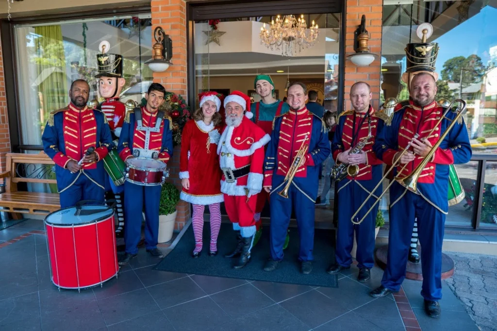 Papai Noel posa sorridente com músicos vestidos como soldados da banda marcial, acompanhado por ajudantes natalinos, em frente à entrada decorada do Hotel Recanto da Serra, uma cena festiva que celebra a magia do Natal com música e encantamento.