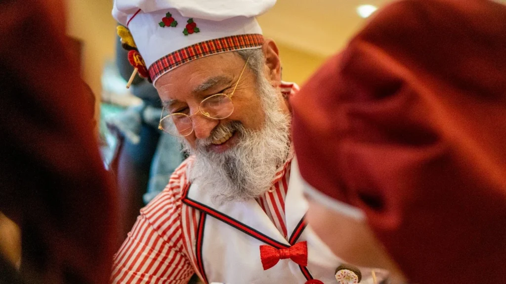 Papai Noel sorridente interage com os hóspedes durante o tradicional café da manhã temático no Hotel Recanto da Serra, usando traje de chef com detalhes natalinos e transmitindo alegria às famílias presentes.