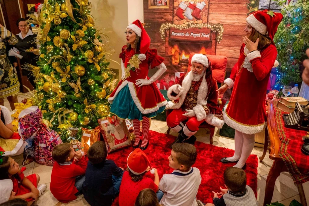 Crianças sentadas ao redor da árvore de Natal assistem à entrega de presentes feita por Papai Noel e ajudantes no Hotel Recanto da Serra, em uma noite mágica com decoração dourada, fantasias natalinas e clima de encantamento.