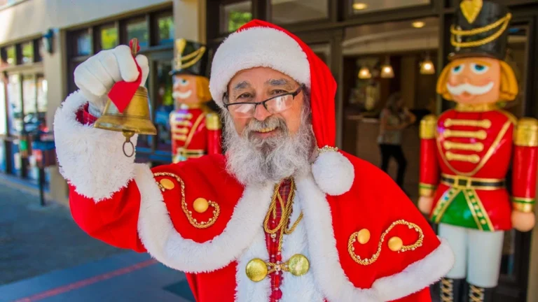 Papai Noel sorridente toca um sino dourado em frente à entrada do Hotel Recanto da Serra, com dois soldadinhos quebra-nozes decorativos ao fundo, anunciando com alegria a chegada do espírito natalino.