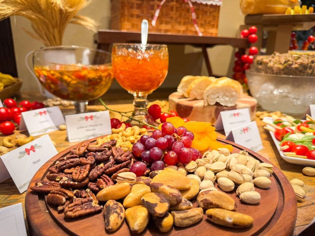 Prato com castanhas, nozes, pistaches, uvas e frutas secas em destaque sobre uma mesa decorada com itens natalinos. Ao fundo, pães, geleias, saladas e aperitivos compõem o buffet de Natal no Hotel Recanto da Serra.