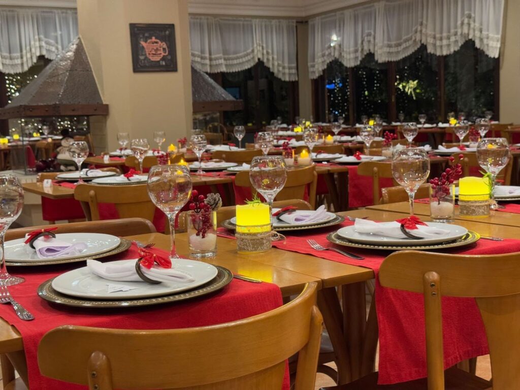 Salão de jantar decorado para o Natal, com mesas arrumadas com toalhas vermelhas, pratos com guardanapos dobrados e taças de vidro. Velas decorativas e arranjos natalinos completam o ambiente aconchegante e elegante do Hotel Recanto da Serra.
