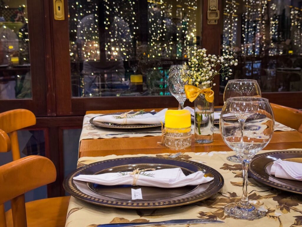 Detalhe de uma mesa elegantemente posta para a ceia de Ano Novo no Hotel Recanto da Serra, com pratos decorados, taças de vidro, vela amarela e arranjo de flores, em frente a janelas iluminadas com luzes festivas ao fundo.