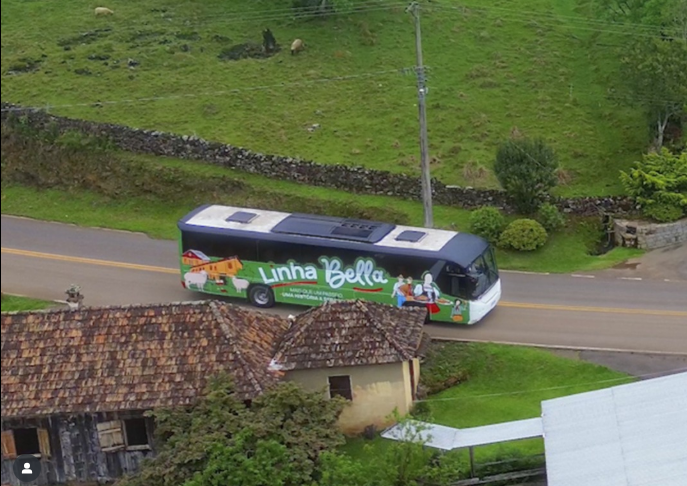 Ônibus do tour Linha Bella circulando por uma estrada rural em Gramado, cercado por paisagens verdes, casas coloniais e áreas típicas da imigração italiana.