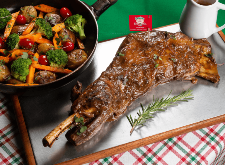 Costela suína suculenta acompanhada de legumes assados, como brócolis, batatas e cenouras, servida com um molho delicioso. A carne é decorada com ramalhete de alecrim, proporcionando um prato saboroso para um jantar em Gramado. O prato é apresentado em uma travessa metálica, com fundo de toalha xadrez.