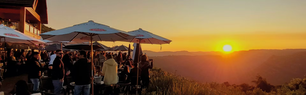 Pôr do sol no Parque Olivas de Gramado, com visitantes reunidos em mesas ao ar livre sob ombrelones, contemplando a vista.
