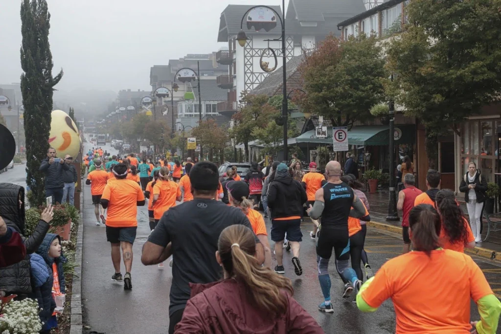 Participantes de uma corrida de rua percorrem a Avenida Borges de Medeiros durante o Choco Esportes, na Choco Páscoa em Gramado. Corredores com camisetas laranja seguem pela via molhada em um dia nublado, enquanto espectadores acompanham nas calçadas, com arquitetura típica da cidade e decoração temática ao longo do percurso.