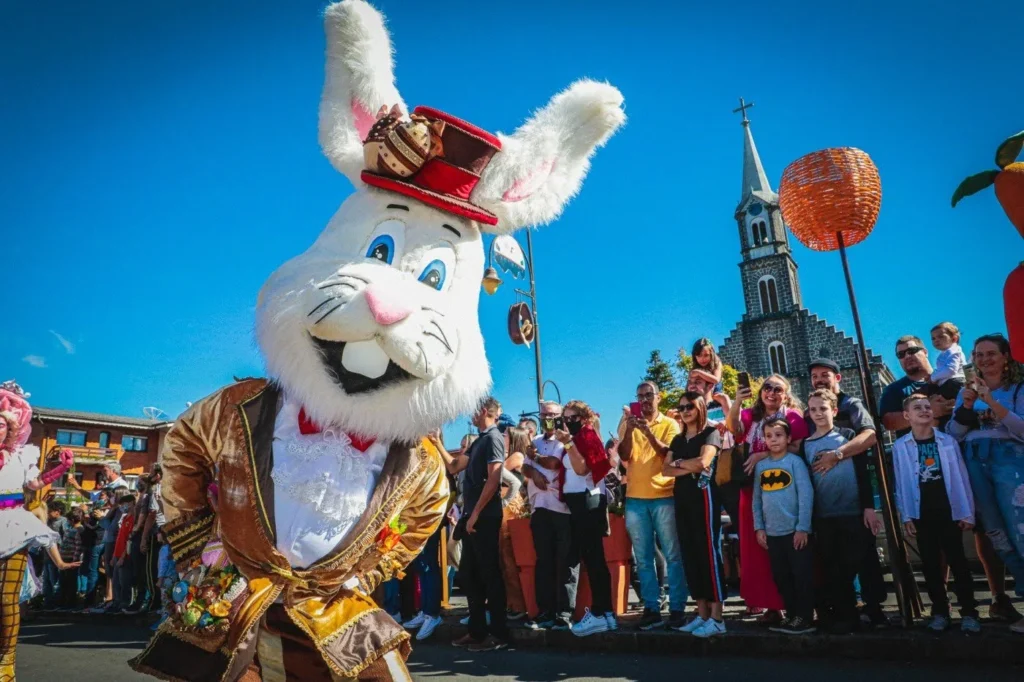 Personagem de coelho gigante e sorridente, vestido com traje elegante e cartola, interagindo com o público durante a Parada de Páscoa na Avenida Borges de Medeiros, em Gramado. Ao redor, famílias e crianças observam e registram o momento, com a Igreja Matriz São Pedro Apóstolo ao fundo e decoração temática colorida sob céu azul, criando um clima festivo e encantador típico do evento.