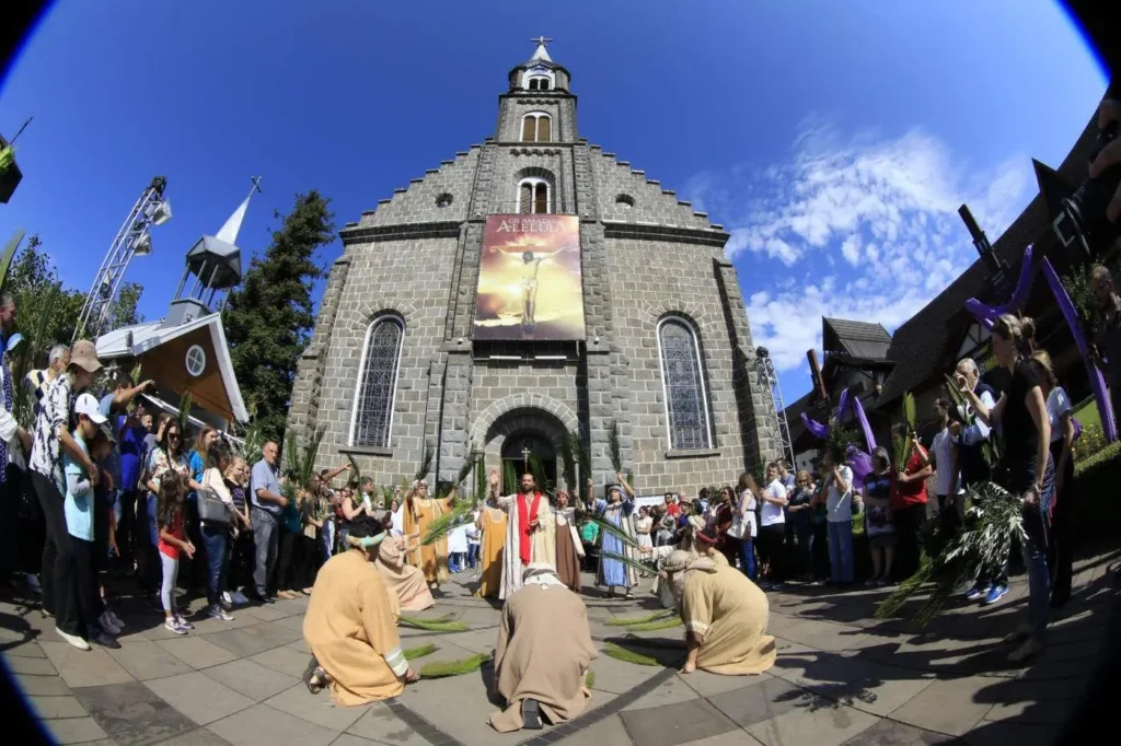 Fiéis participam da Procissão de Ramos em frente à Igreja Matriz São Pedro Apóstolo, em Gramado, marcando a abertura do evento Gramado Aleluia. No centro, encenação religiosa com personagens bíblicos, enquanto o público ao redor acompanha segurando ramos, sob céu azul, criando um ambiente solene e simbólico da celebração.