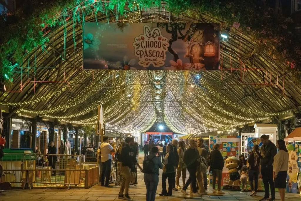 Movimento de visitantes na Rua Coberta durante a Choco Páscoa em Gramado, com estrutura decorada por arcos de madeira, iluminação com milhares de luzes pendentes e um letreiro do evento ao centro. Pessoas circulam entre espaços temáticos e atrações sob o teto iluminado, criando um ambiente acolhedor, festivo e encantador à noite.