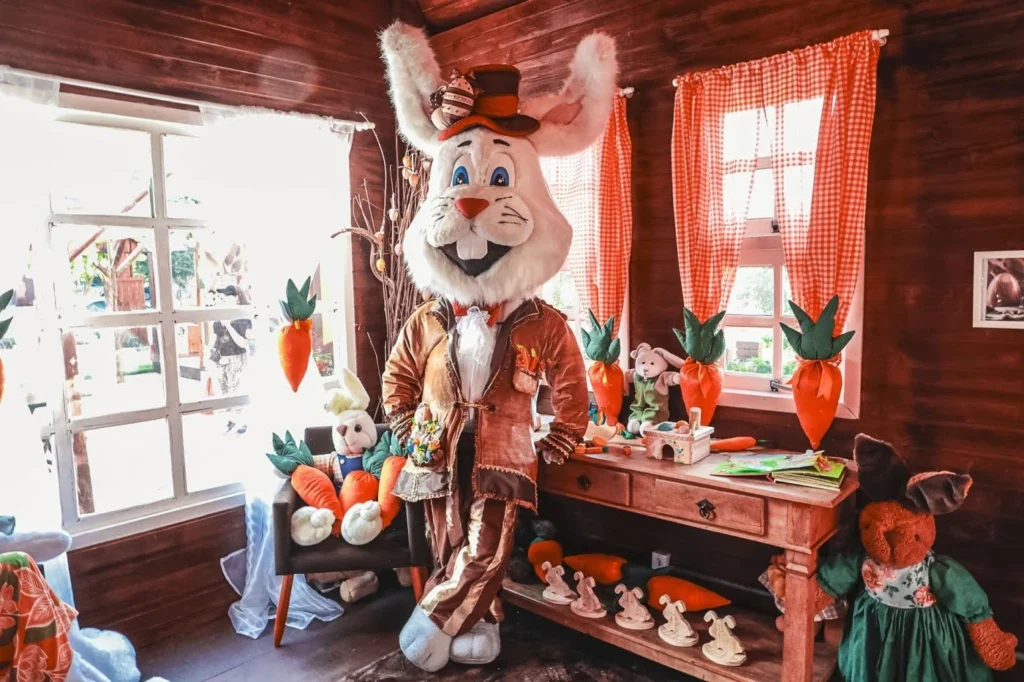 Interior da Casa do Pascoalino na Choco Páscoa em Gramado, com um personagem de coelho em tamanho real vestido com roupas elegantes e cartola, posicionado em um ambiente rústico de madeira. O espaço é decorado com cenouras coloridas, pelúcias de coelhos e brinquedos sobre uma mesa e poltrona, além de janelas com cortinas xadrez laranja que deixam a luz natural entrar, criando uma atmosfera acolhedora, lúdica e temática de Páscoa.