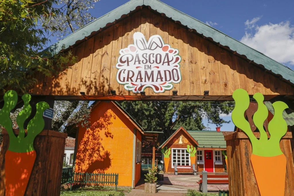 Entrada decorativa do evento “Páscoa em Gramado”, com portal de madeira rústica exibindo o nome do festival, enfeitado com ilustrações de coelhos e cenouras em cores vibrantes. Ao fundo, pequenas casinhas coloridas e charmosas, lembrando um vilarejo temático, sob céu azul e cercado por árvores, transmitindo um clima lúdico e festivo típico da Choco Páscoa em Gramado.