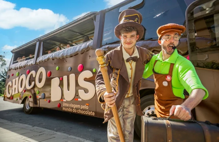 Personagens caracterizados recebem visitantes em frente ao Chocobus, ônibus temático em Gramado decorado com elementos de chocolate e cores vibrantes. A atração oferece uma experiência interativa e divertida pelo universo do chocolate, com participantes visíveis no interior do veículo.