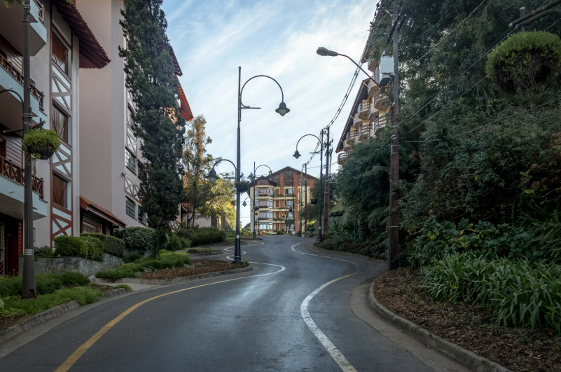 Uma rua sinuosa e charmosa no centro de Gramado, ladeada por árvores altas e prédios em estilo arquitetônico europeu (enxaimel) com detalhes em madeira. A via apresenta curvas suaves demarcadas por faixas no asfalto, luminárias clássicas de ferro e canteiros floridos, transmitindo a atmosfera acolhedora e a sofisticação cultural da cidade gaúcha próxima ao Hotel Recanto da Serra.
